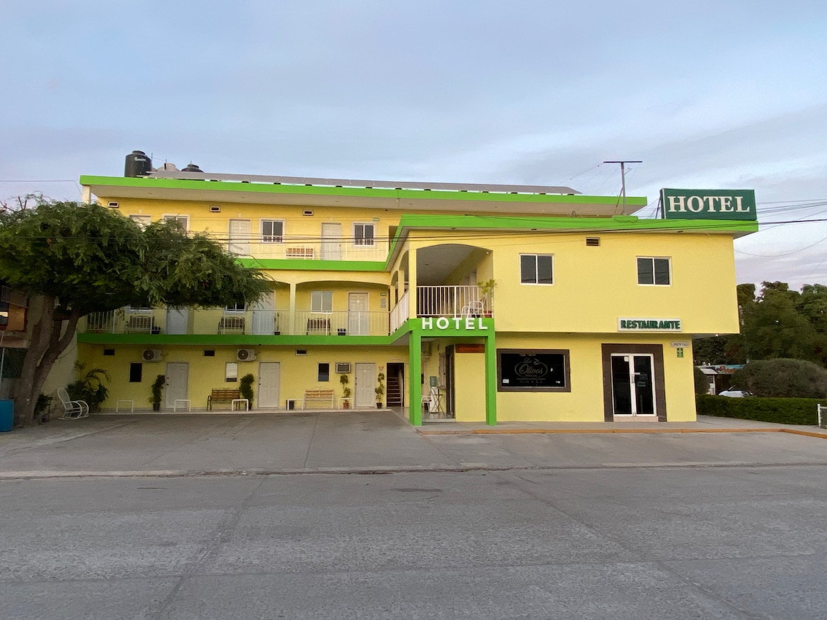 A bright yellow hotel building features a green roof and signage. The ground floor includes a restaurant area with large windows, while balconies are visible on the upper level. Outdoor seating is arranged in a welcoming manner, and parking is available in front.