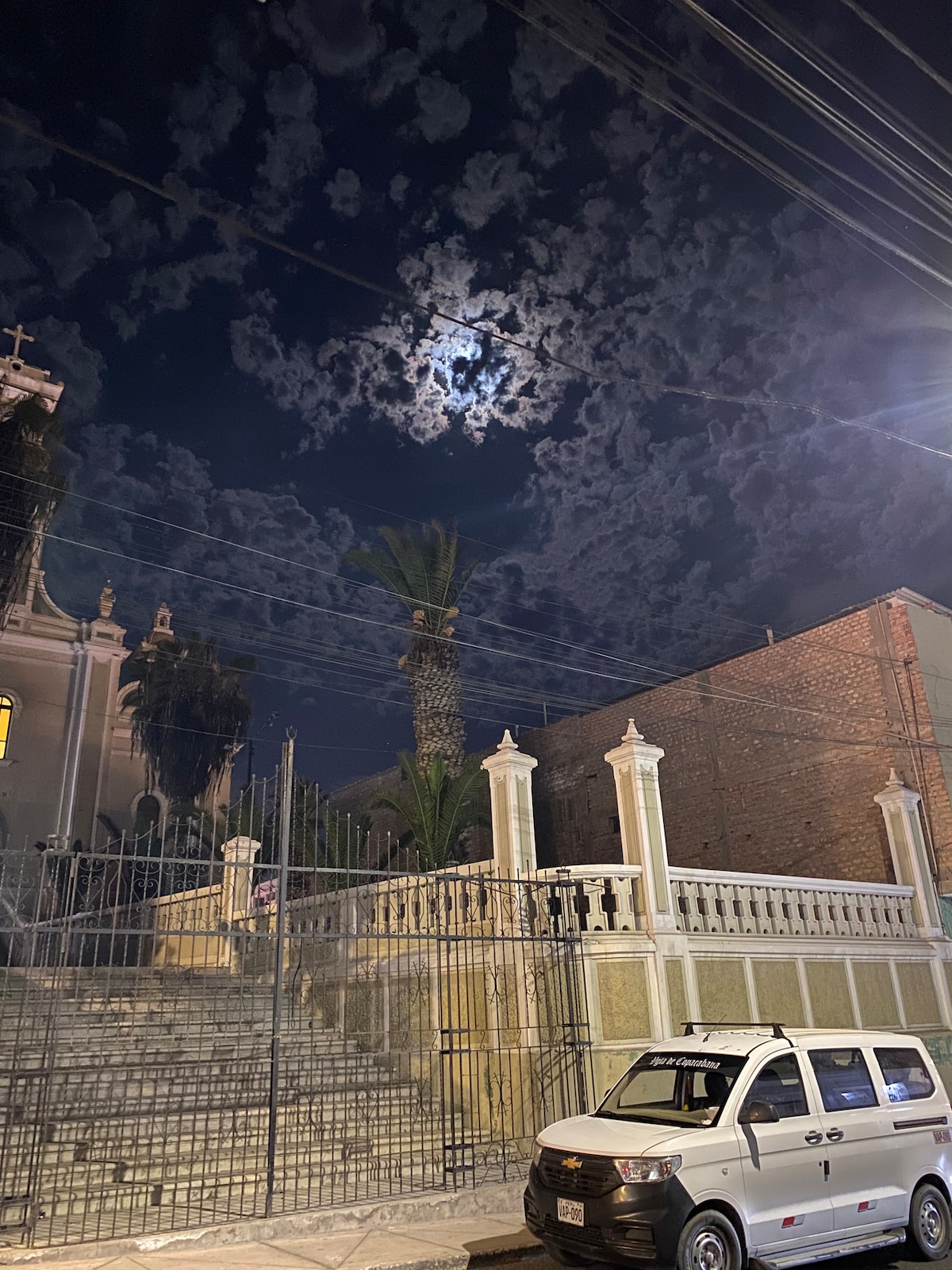 A moonlit night scene features a prominent building with ornate architectural details. Clouds drift across the sky, and a glowing full moon can be seen peeking through. Palm trees are positioned adjacent to the structure, while a parked vehicle is visible in the foreground.