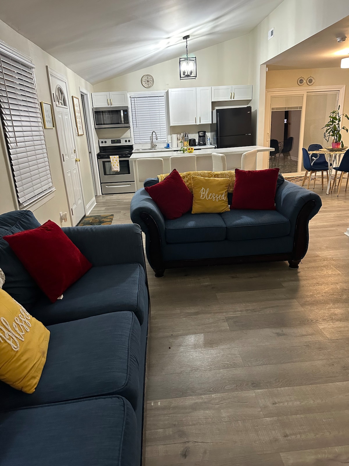 A comfortable living area features a blue sofa and loveseat, adorned with decorative pillows in red and yellow. Natural light filters through the window blinds, illuminating the space. An open kitchen layout is visible in the background, emphasizing an inviting atmosphere.