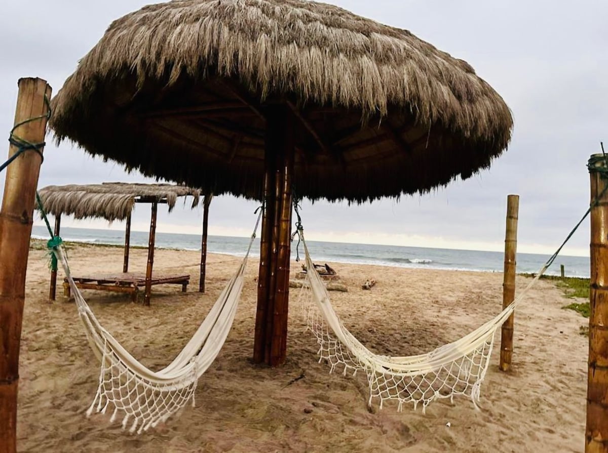 Two hammocks are suspended under a large thatched gazebo, set on a sandy beach. The ocean is visible in the background, with gentle waves lapping at the shore. A soft, overcast sky complements the serene coastal setting.