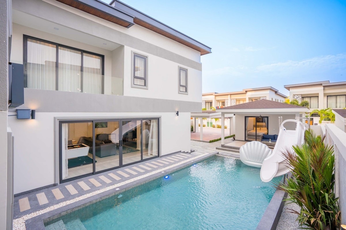 The exterior view showcases a multi-level villa with large glass doors leading to a pool area. A water slide is visible next to the pool, surrounded by modern landscaping. The structure features a mix of neutral tones and contemporary architectural elements.