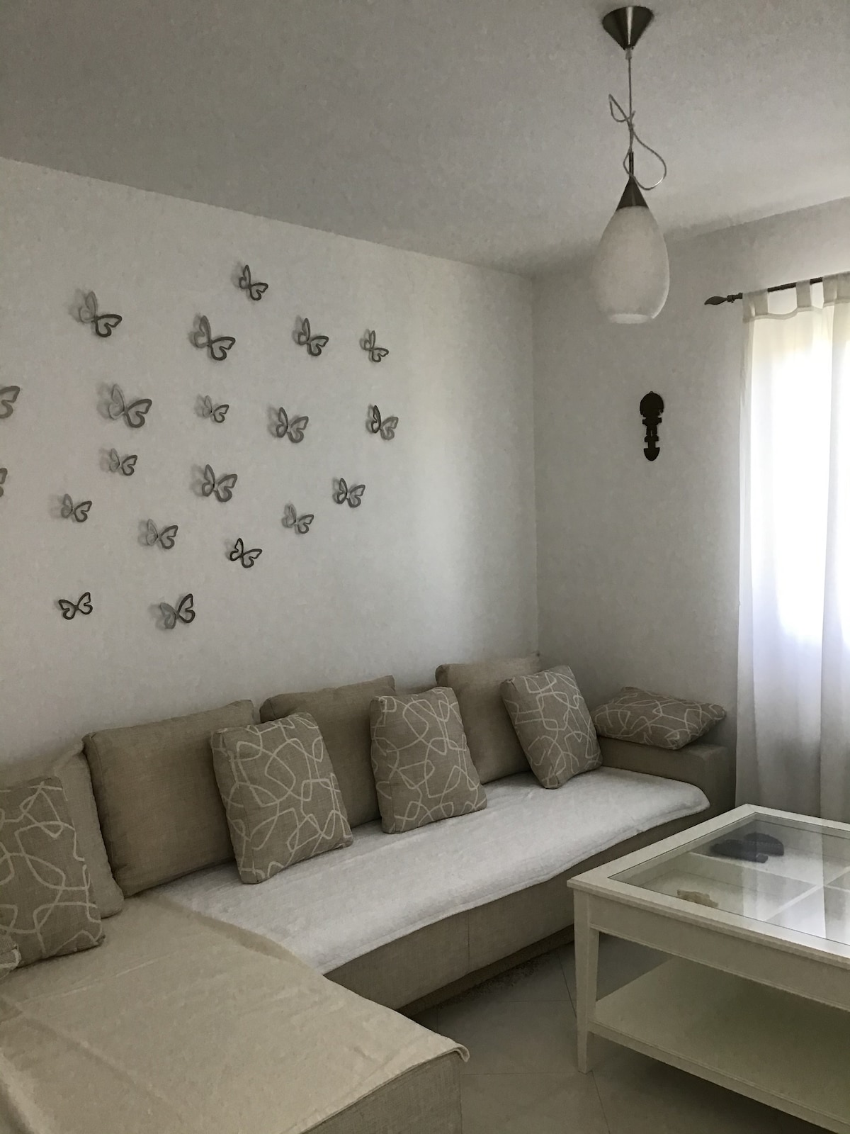 A light-filled living area features a spacious beige sofa adorned with patterned cushions. A glass coffee table sits at the center, and a wall is decorated with an arrangement of butterfly art. A soft curtain allows natural light to filter in.