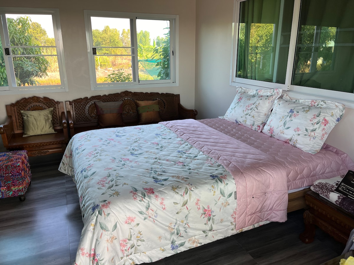A spacious bedroom features a large bed with floral bedding and decorative pillows. Natural light enters through multiple windows, illuminating the room. A wooden seating area is arranged to the side, enhancing the comfortable and relaxing environment.