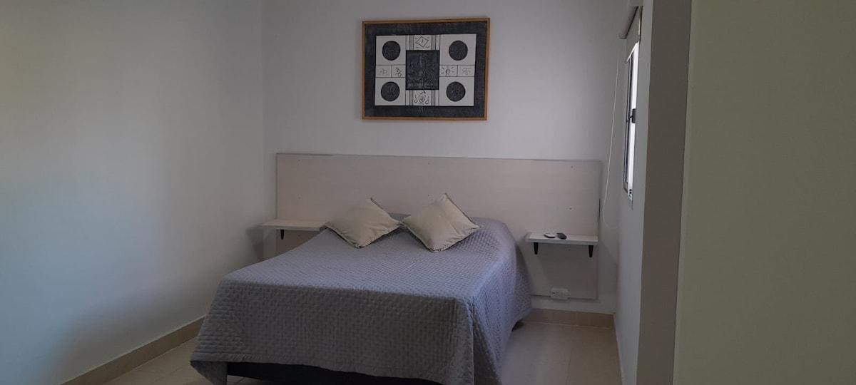 A well-lit sleeping area is presented with a spacious bed, dressed in a neutral-colored coverlet. Two decorative pillows are placed against a wooden headboard. Light enters through a nearby window, complementing the minimalist design of the room.