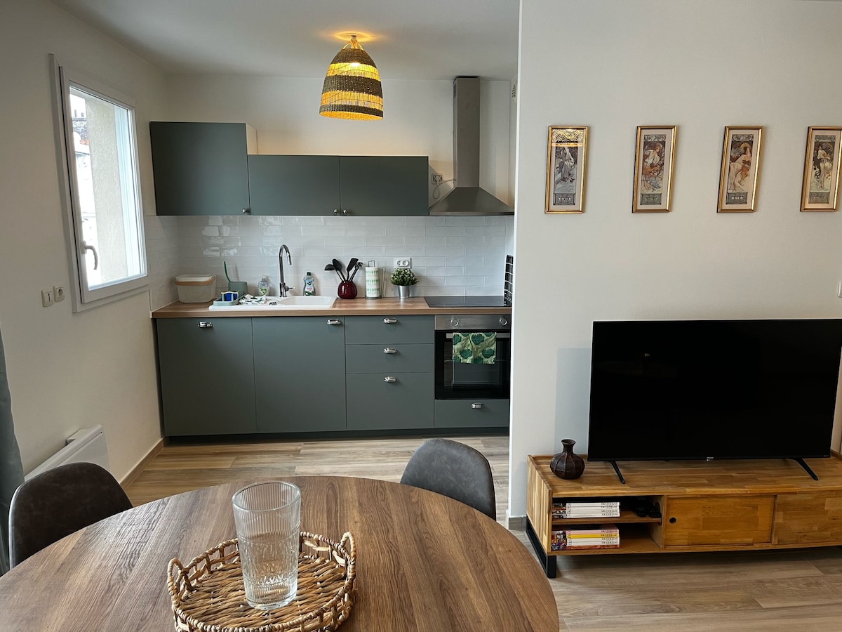 The kitchen features modern cabinetry in a muted green tone, with stainless steel appliances and ample counter space. A round dining table is positioned nearby, accompanied by grey upholstered chairs. A television is displayed on a wooden media console, and decorative wall art adds character.