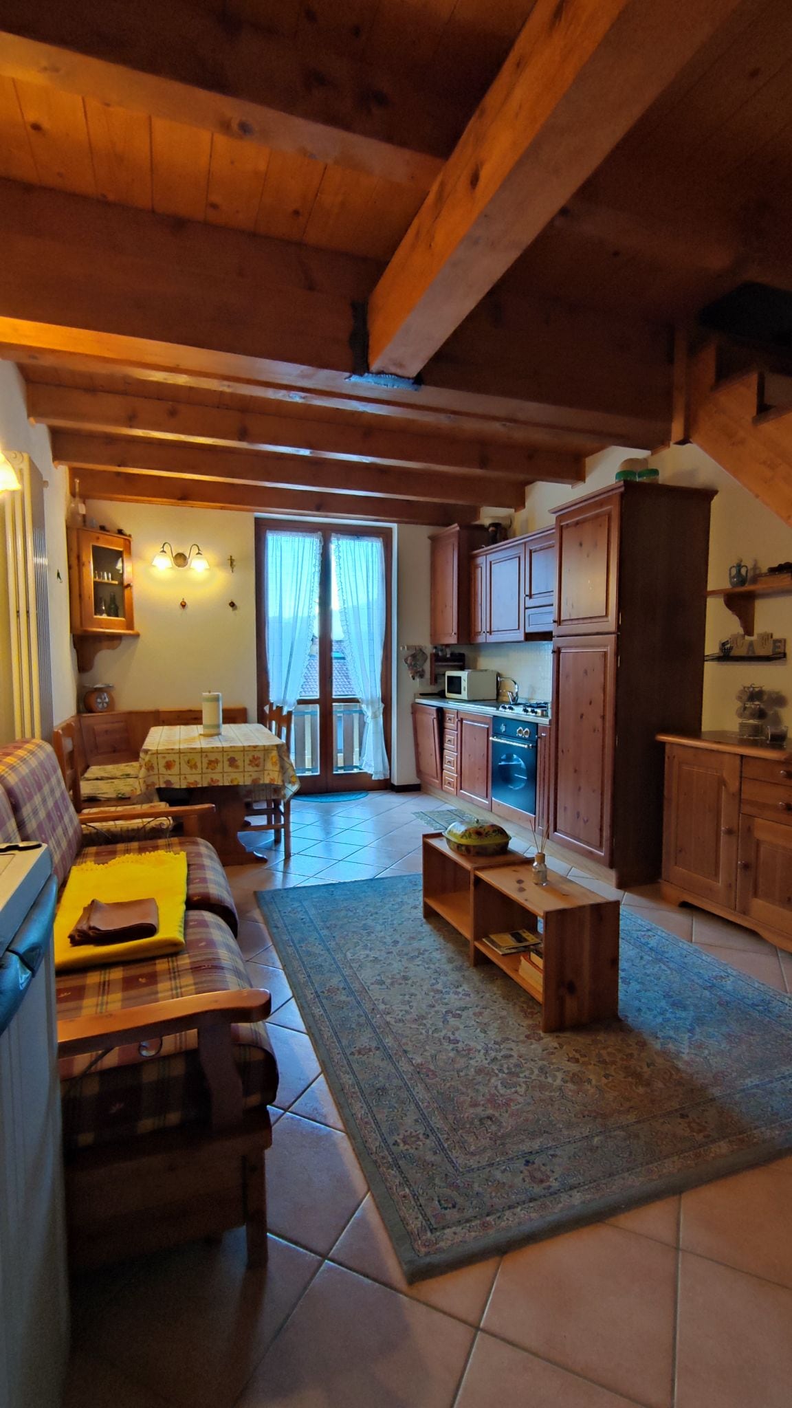A comfortable living space features wooden beams and warm cabinetry. A cozy seating area with a patterned rug sits opposite a dining table. Natural light filters through the window dressed in light curtains, while a small table displays decorative items. An inviting atmosphere is evident.