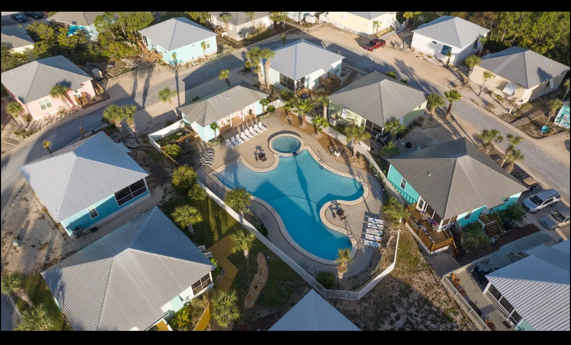 An aerial view showcases a well-maintained community featuring several colorful cottages surrounding a central swimming pool. Lush greenery and palm trees offer a tropical feel, while the pool area includes lounge chairs and an inviting circular shape, ideal for guest relaxation.