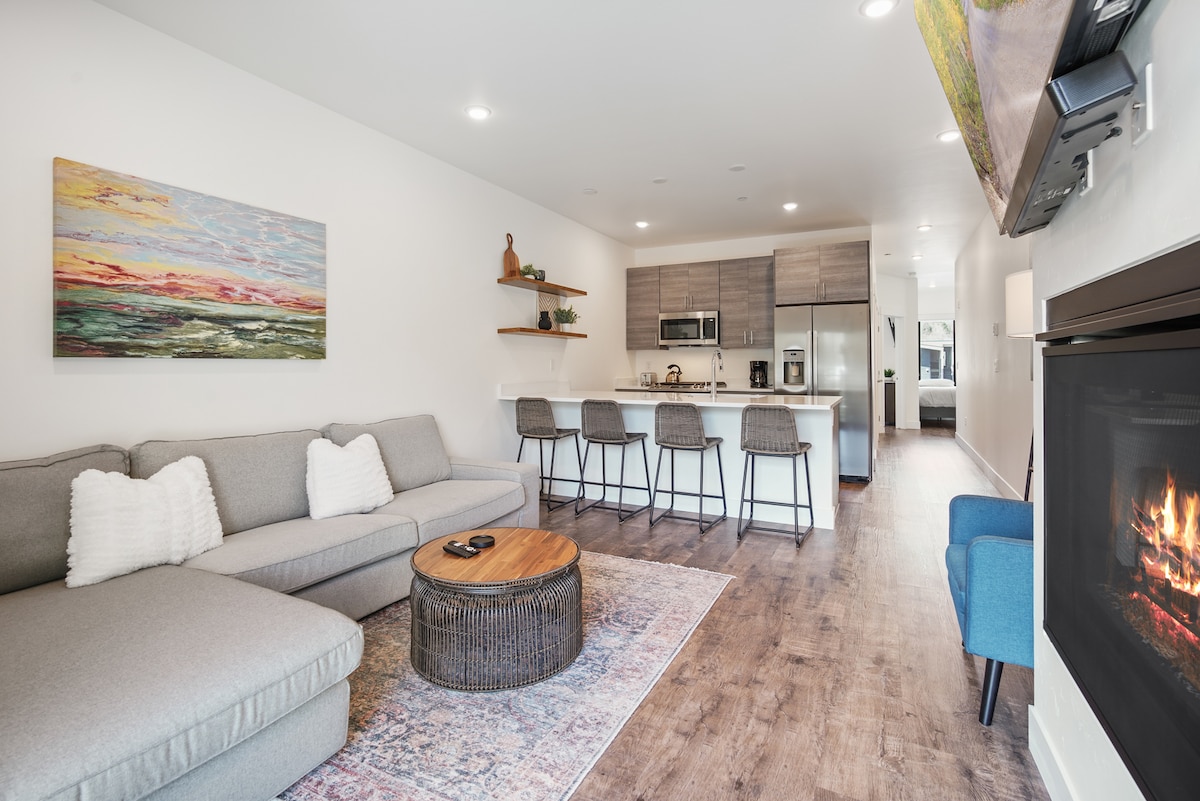 A spacious living area features a modern sectional sofa with decorative pillows, a round coffee table, and a cozy fireplace. The open layout includes a dining space with three barstools, leading to a well-equipped kitchen that showcases sleek cabinetry and stainless-steel appliances.