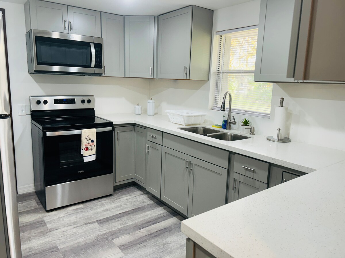 The kitchen features modern gray cabinets complemented by a stainless-steel refrigerator and oven. A spacious countertop extends across one side, with a double sink positioned under a window. Natural light enhances the well-organized space, which includes essential appliances for meal preparation.