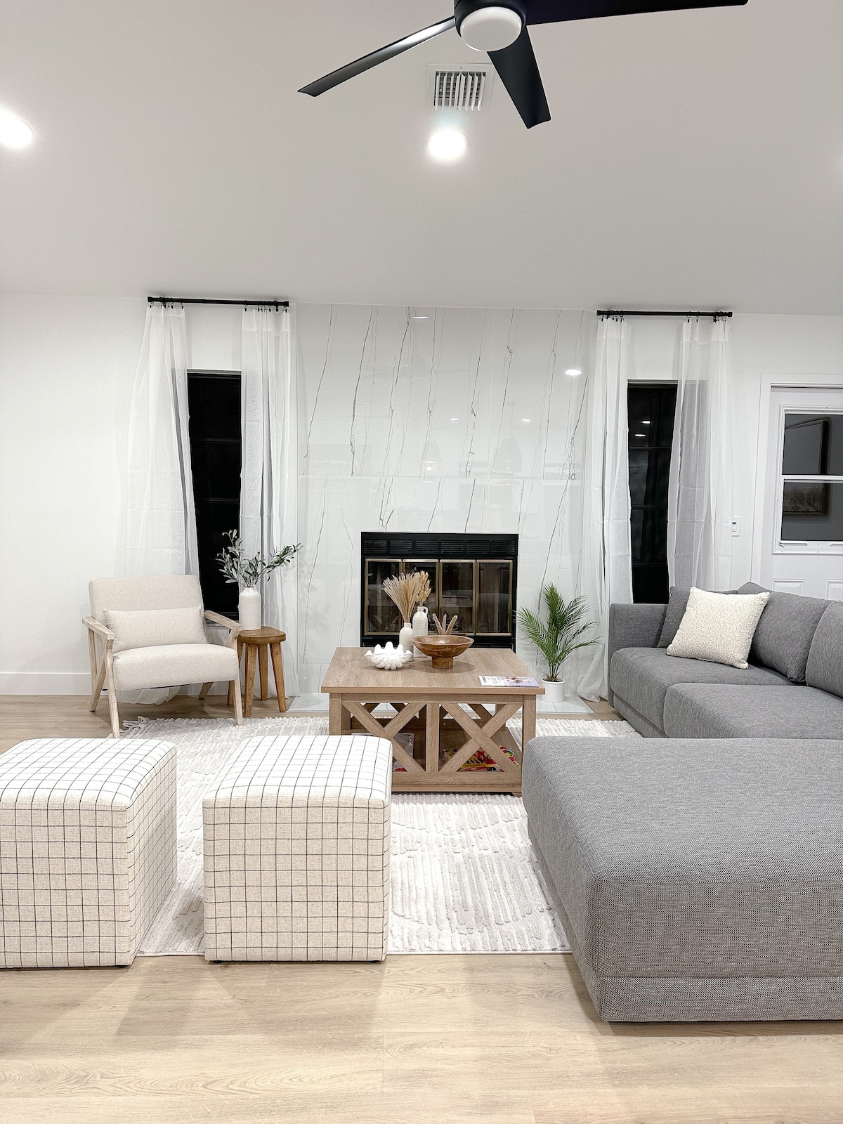 The living area features a cozy arrangement with a gray sectional sofa and a wooden coffee table at the center. Two patterned ottomans are positioned nearby. A fireplace is set against a white backdrop, flanked by large windows dressed with sheer curtains.