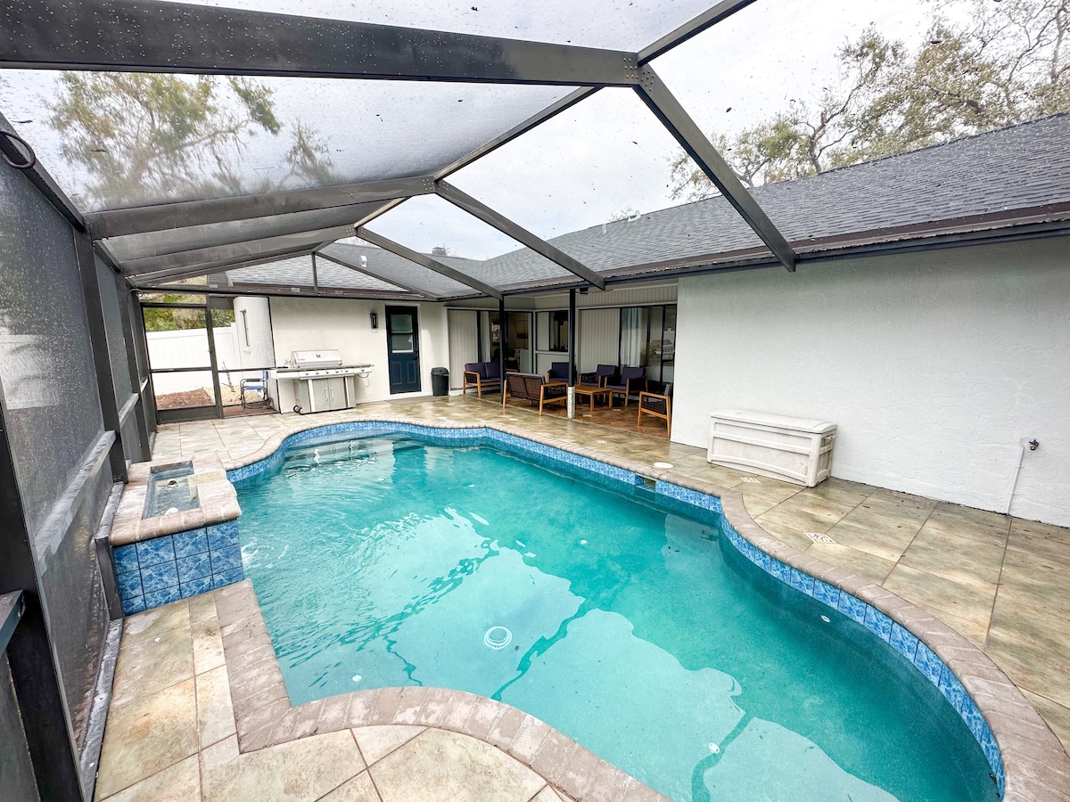 A private swimming pool is surrounded by a screened patio, featuring a relaxing seating area. The pool exhibits a curved design with clear water, while the surrounding tiled deck is complemented by a shaded area and an outdoor dining space in the background.
