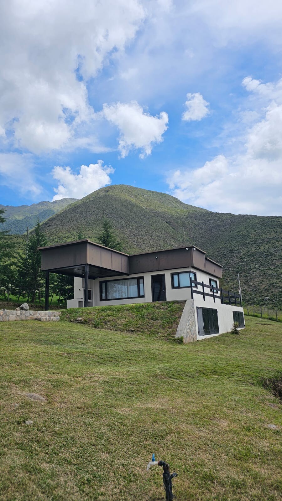 A modern house is situated on a grassy terrain, surrounded by picturesque mountains. Large windows provide scenic views of the landscape, while the building's unique architectural design features a mix of dark and light tones. The outdoor area showcases natural greenery.