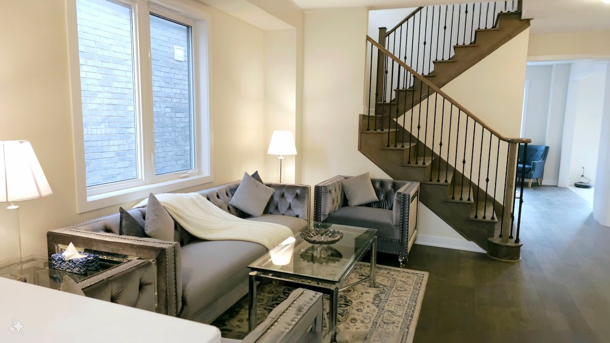 A modern living area features a plush grey sectional sofa adorned with a soft blanket, accompanied by a glass coffee table. Well-placed lamps illuminate the space, while large windows provide ample natural light. The upscale staircase is visible in the background, enhancing the room's open layout.