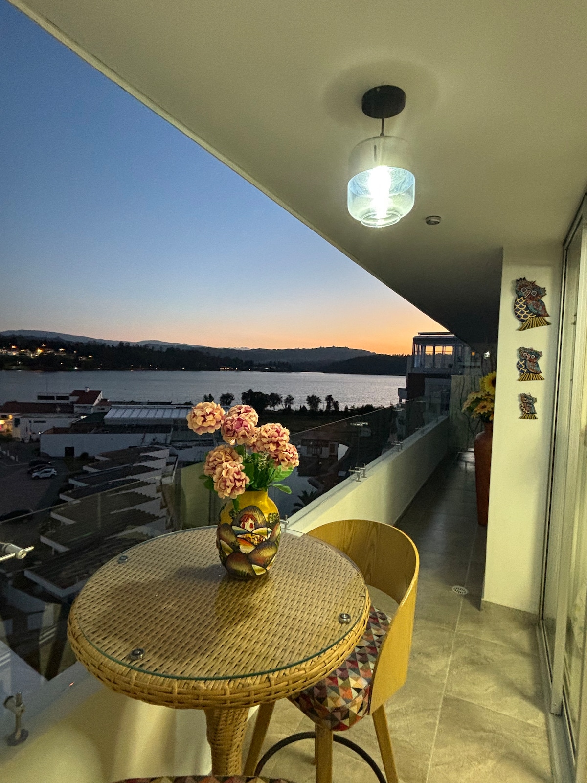 A balcony features a round table with a woven surface and two wooden chairs, accompanied by a vase of fresh flowers. The scenic view of the lake is highlighted against a dusky sky as twilight approaches, enhancing the tranquil ambiance of the space.