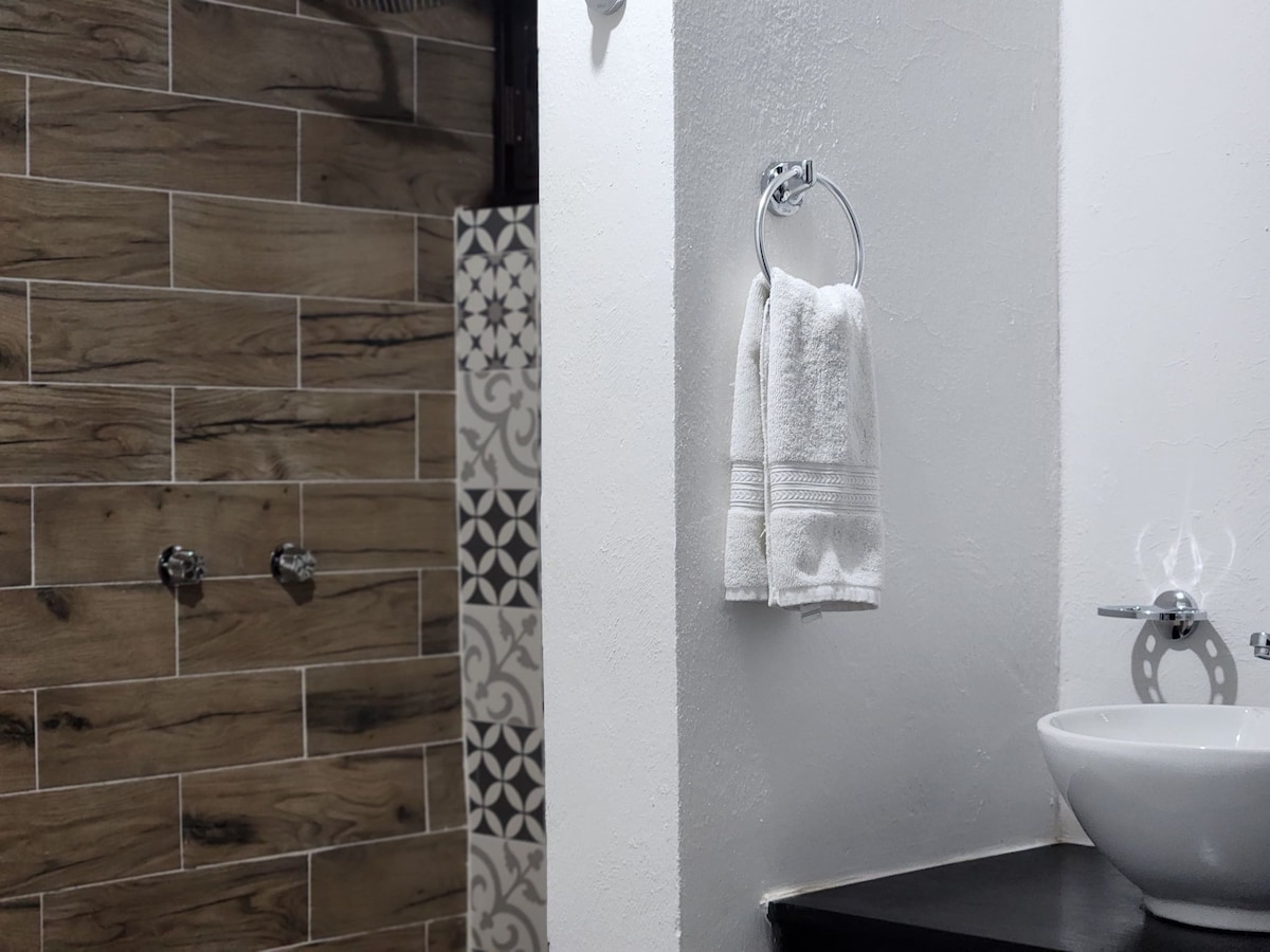 A modern bathroom is displayed, featuring a sleek white sink with a chrome faucet. A hand towel is neatly arranged on a chrome ring. The walls are adorned with textured tiles, combining wood-like and patterned designs, providing a clean and contemporary feel.