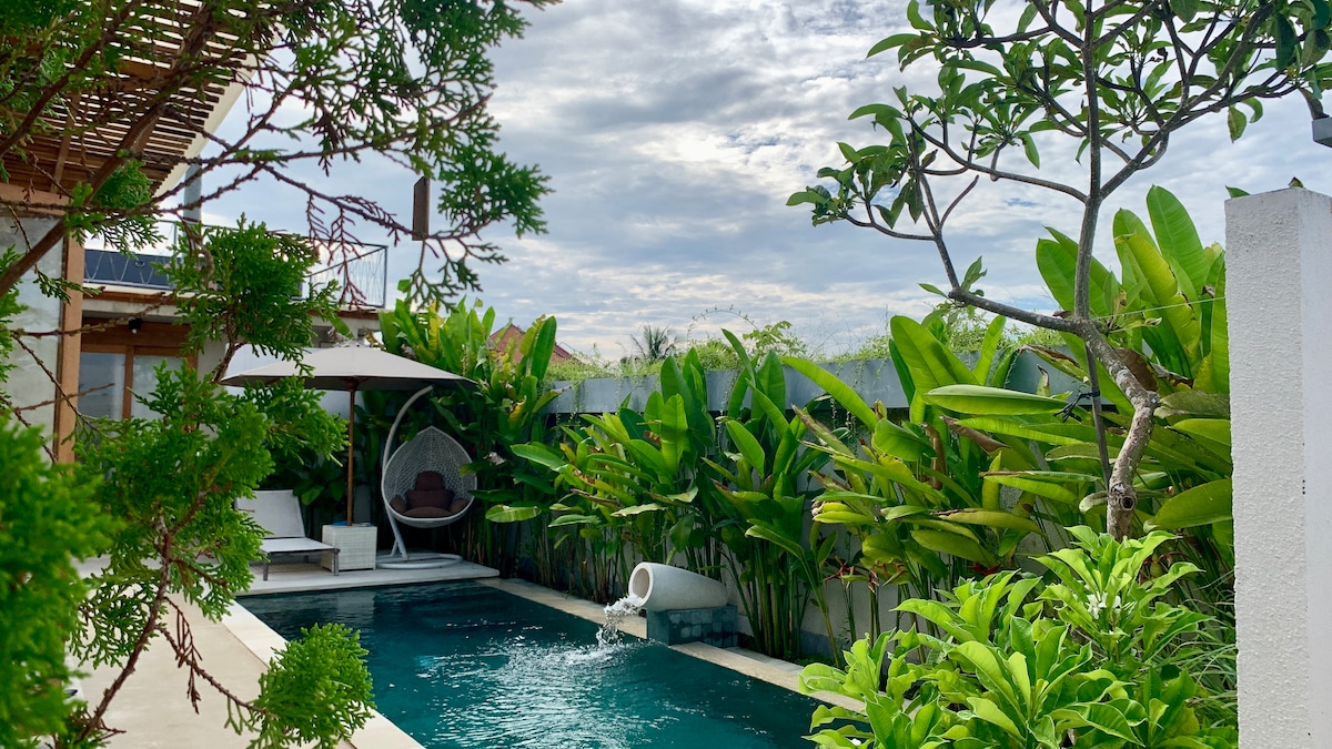 A private pool is surrounded by lush greenery, featuring tropical plants and trees. A shaded lounge area includes a cozy hanging chair, inviting relaxation. The pool area is designed for tranquility, blending seamlessly into the natural environment.