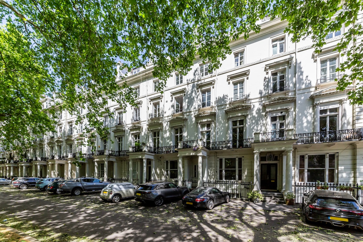 An elegant row of white-columned buildings is lined with leafy trees, creating a shaded atmosphere. Cars are parked along the street, while balconies adorn the upper levels, enhancing the architectural appeal of the neighborhood.
