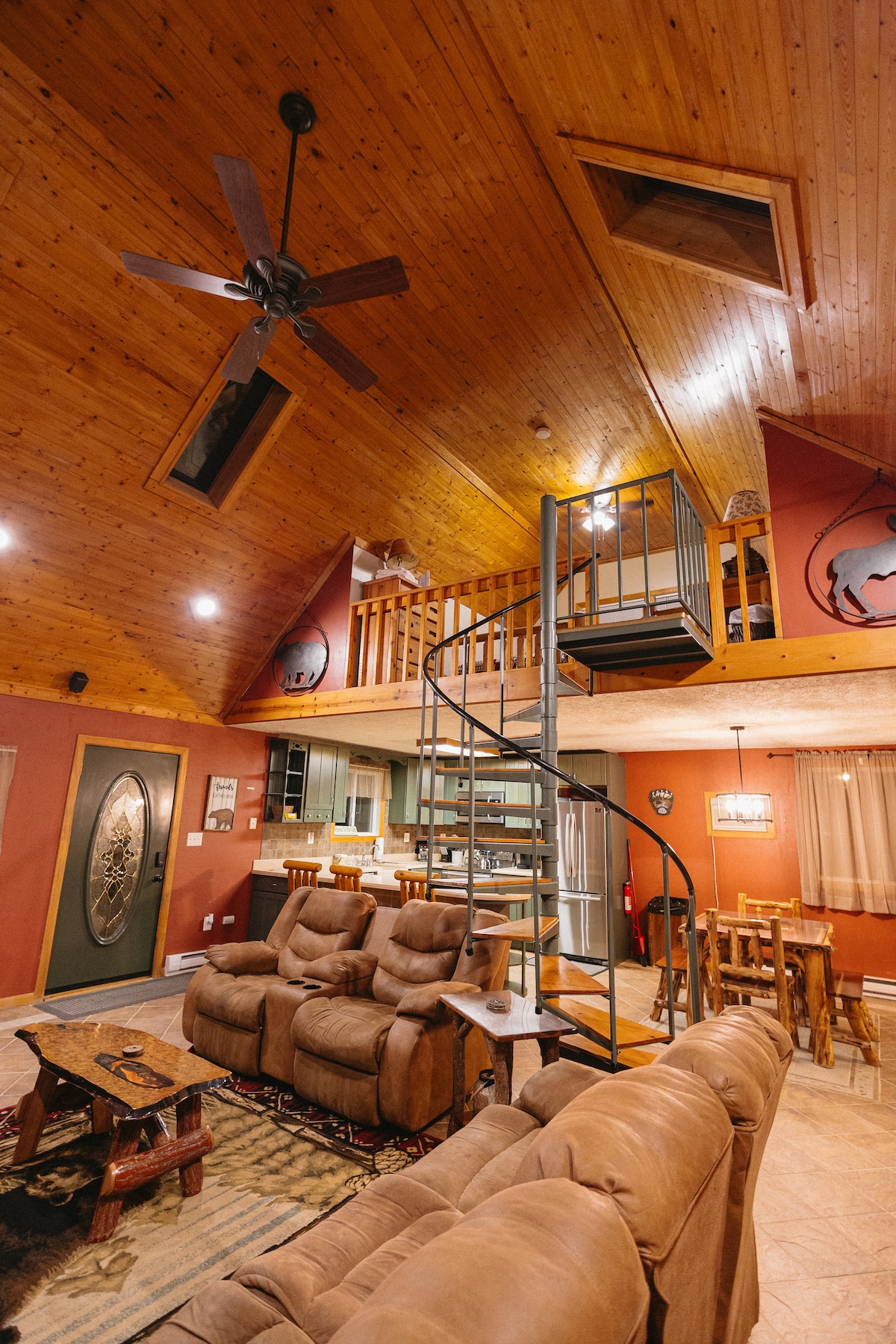 The interior features a spacious open floor plan with a spiral staircase leading to a loft. Cathedral ceilings adorned with wooden accents create an inviting atmosphere. A comfortable seating area with brown couches is visible, along with a dining area and well-equipped kitchen in the background.