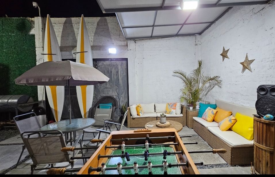 An outdoor lounge area is furnished with a sectional sofa adorned with colorful cushions and a glass table. A foosball table occupies the space, while an umbrella offers shade. Decor elements include potted plants and wall accents, creating a relaxed environment.