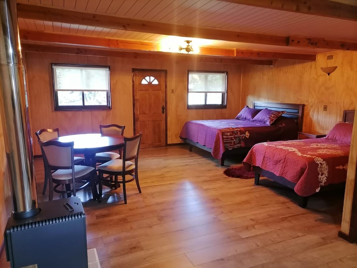 A spacious studio layout is presented, featuring two beds dressed in vibrant red linens. A round dining table with four chairs is positioned near the entrance, allowing for comfortable seating. Natural light filters through windows, and a wood stove adds to the cozy ambiance.
