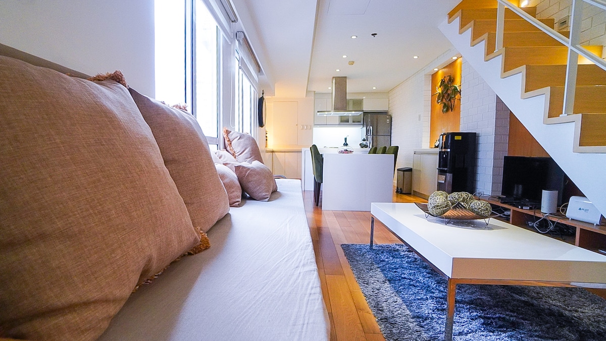 A spacious living area is highlighted by plush cushions on a light-colored sofa, facing a sleek coffee table. Natural light floods through large windows, illuminating the open layout that transitions to a modern kitchen in the background, with a contemporary staircase visible to the side.