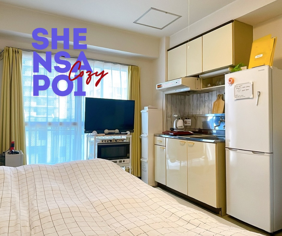 A well-arranged studio apartment showcases a spacious bed in the foreground. Behind it, a kitchenette is equipped with a refrigerator and a stove. A television is positioned across from the bed, and light filters in through large windows with sheer curtains.