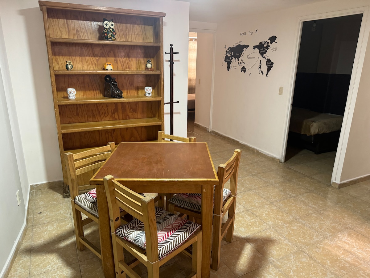 A wooden dining table with four matching chairs is positioned in a bright room. A shelving unit stands against the wall, displaying decorative items and utensils. The space is well-lit, offering a view to another room in the background.