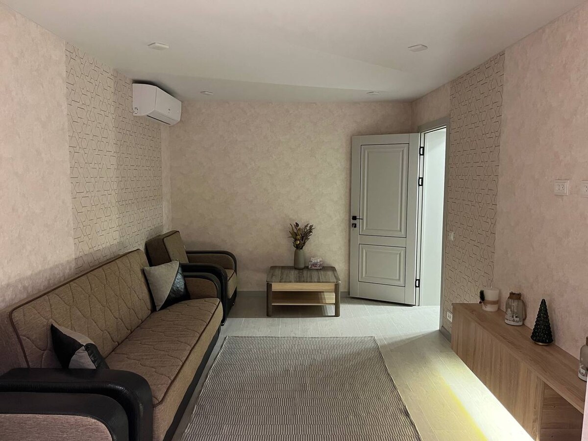 A modern living area is furnished with a neutral-colored sofa and a coffee table. The walls are adorned with a subtle patterned wallpaper, and ample natural light illuminates the room. A door leads to another space, while a simple decor piece sits on the sideboard.