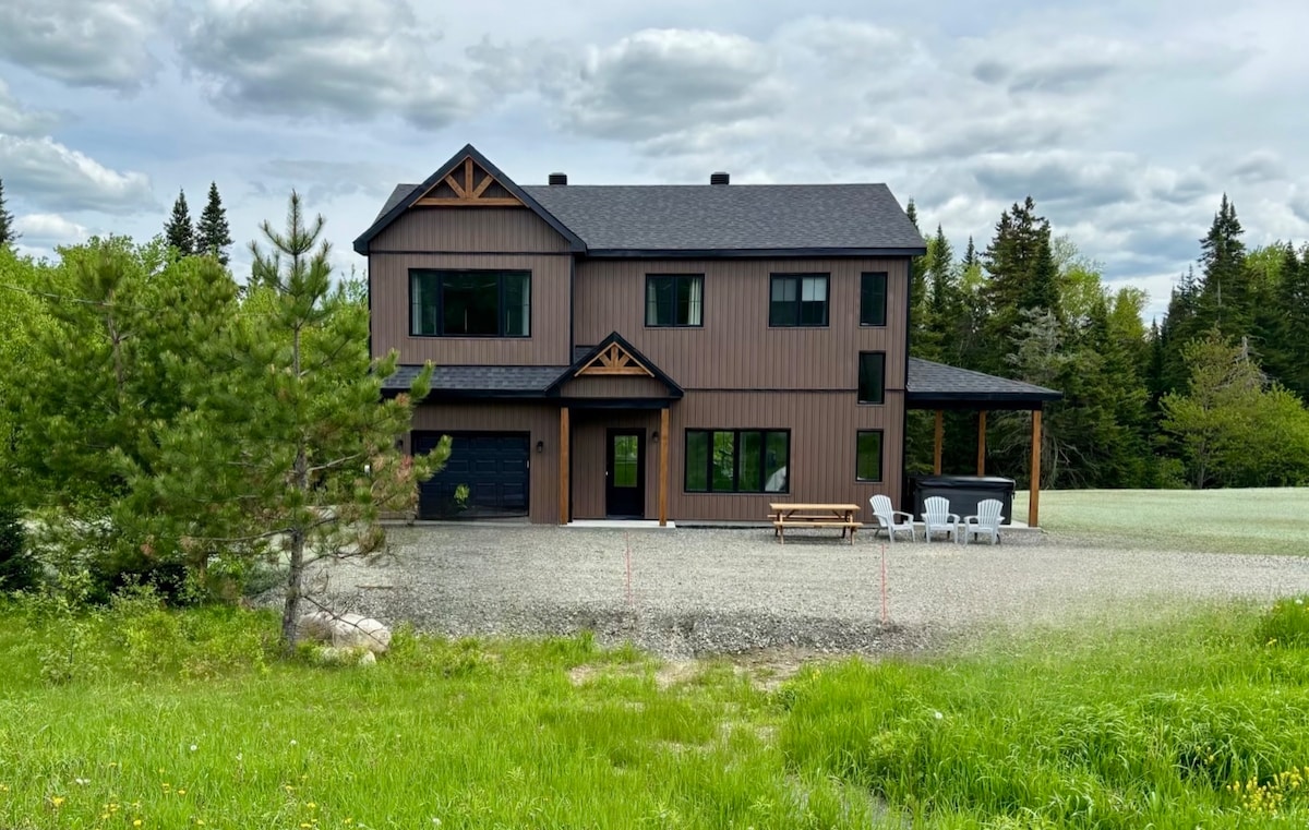 A modern two-story chalet is nestled among trees, featuring a combination of wood and dark finishes. Large windows enhance natural light, while a picnic table sits on the gravel path in front. Lawn areas with greenery extend to the horizon, framed by a cloudy sky.