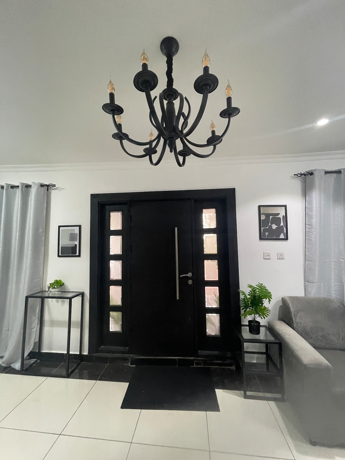 A well-lit entryway features a contemporary chandelier hanging from the ceiling. A black front door with rectangular glass panels stands at the center. Grey curtains frame the sides, while a sleek table and a small potted plant are positioned nearby on the polished floor.