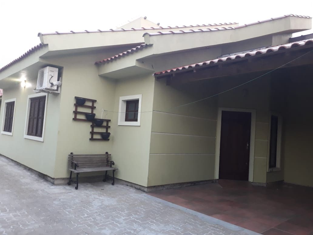 A green house exterior is framed by a sloped roof with terracotta tiles. A wooden bench is positioned beside the entrance. An air conditioning unit is mounted on the wall, and several windows add natural light to the facade.