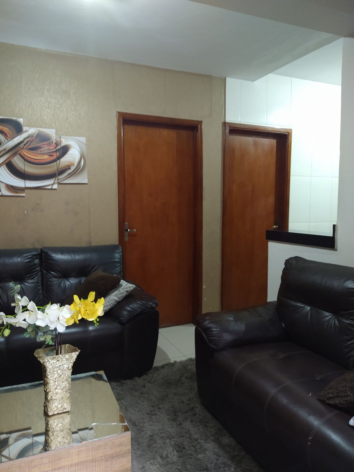 A cozy living area is furnished with dark leather sofas positioned around a glass coffee table. A decorative vase with flowers adds a touch of color. Two wooden doors are visible in the background, leading to other rooms, against a neutral-toned wall.