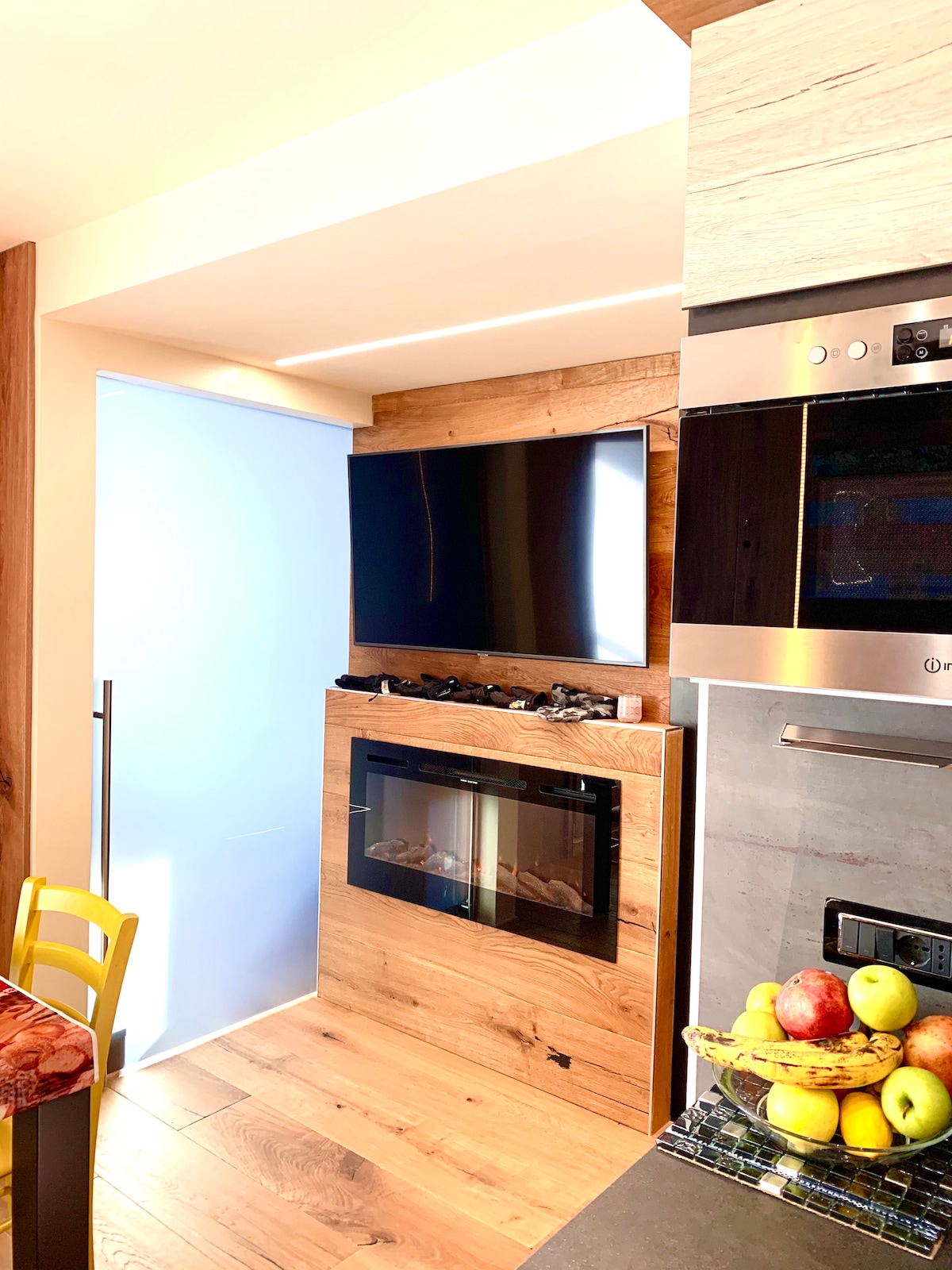 A modern kitchenette features a sleek television mounted above a contemporary electric fireplace. Textured wooden accents provide warmth to the space. A refrigerator is discreetly integrated into the cabinetry, and a bowl of fresh fruit adds a touch of color to the countertop.