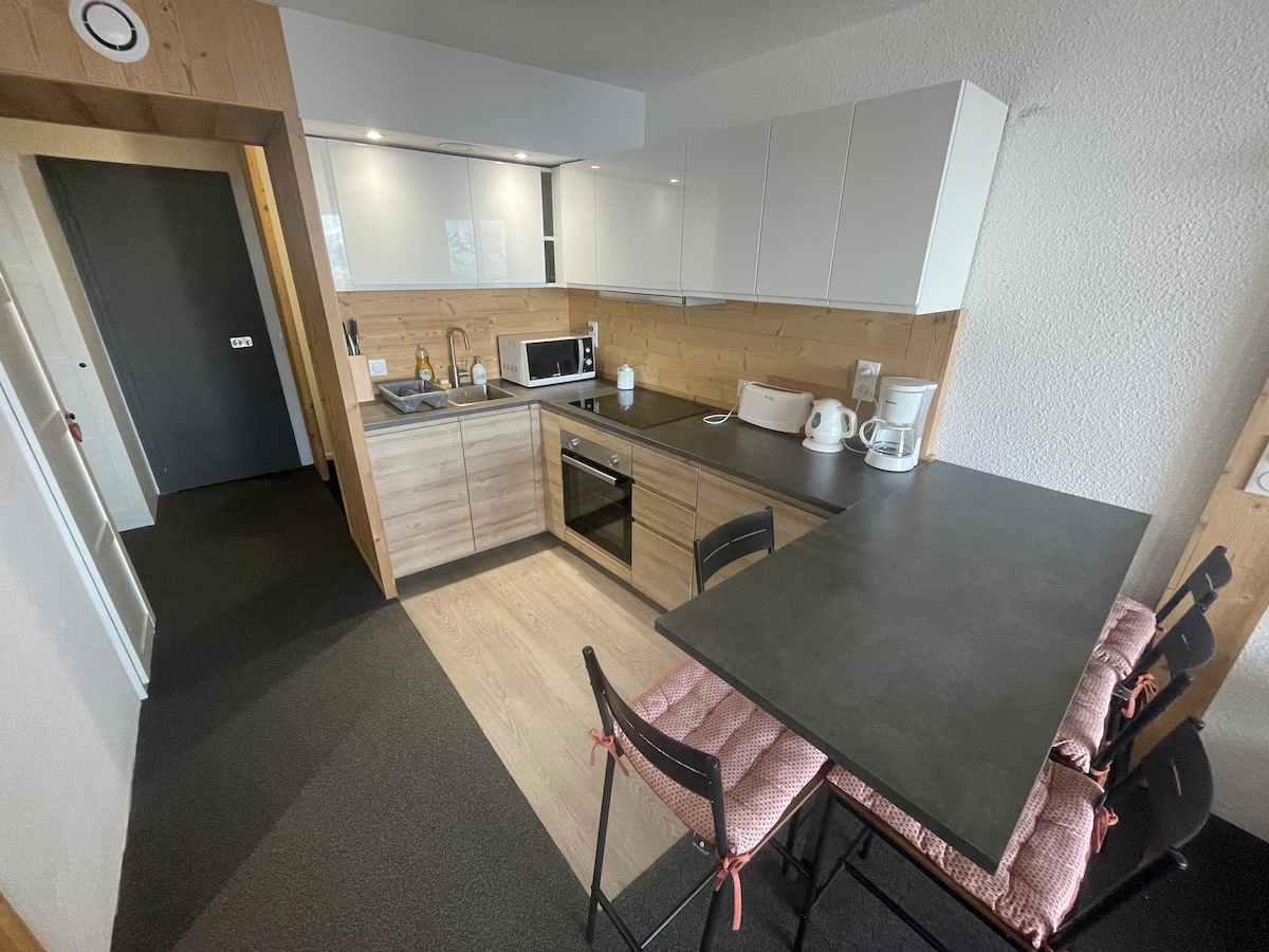 A modern kitchen space is showcased, featuring wood cabinetry and sleek white countertops. Essential appliances including a microwave and kettle are visible. A compact dining table with chairs is positioned near the entrance, providing a cozy spot for meals. The floor exhibits a neutral tone.