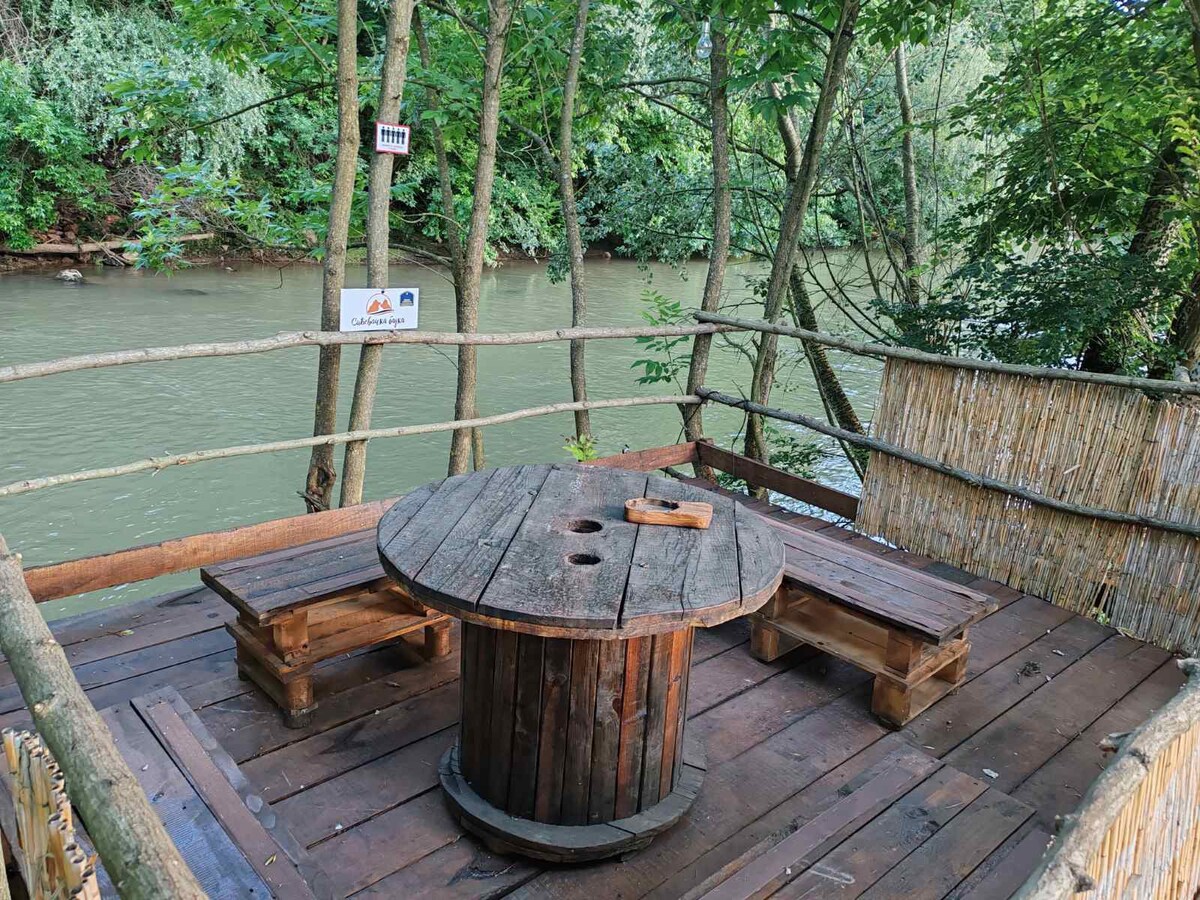 A wooden deck is positioned by the river, featuring a circular table complemented by two benches. Surrounded by trees, the area offers a serene setting with views of the water. Two signs are visible on the nearby tree and fencing.