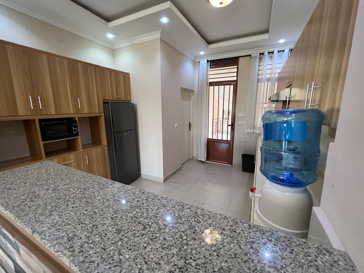 A modern kitchen features wooden cabinetry and a granite countertop. A refrigerator and microwave are visible, along with a water cooler nearby. Natural light enters through a door leading outside, while tiles cover the floor for easy maintenance.