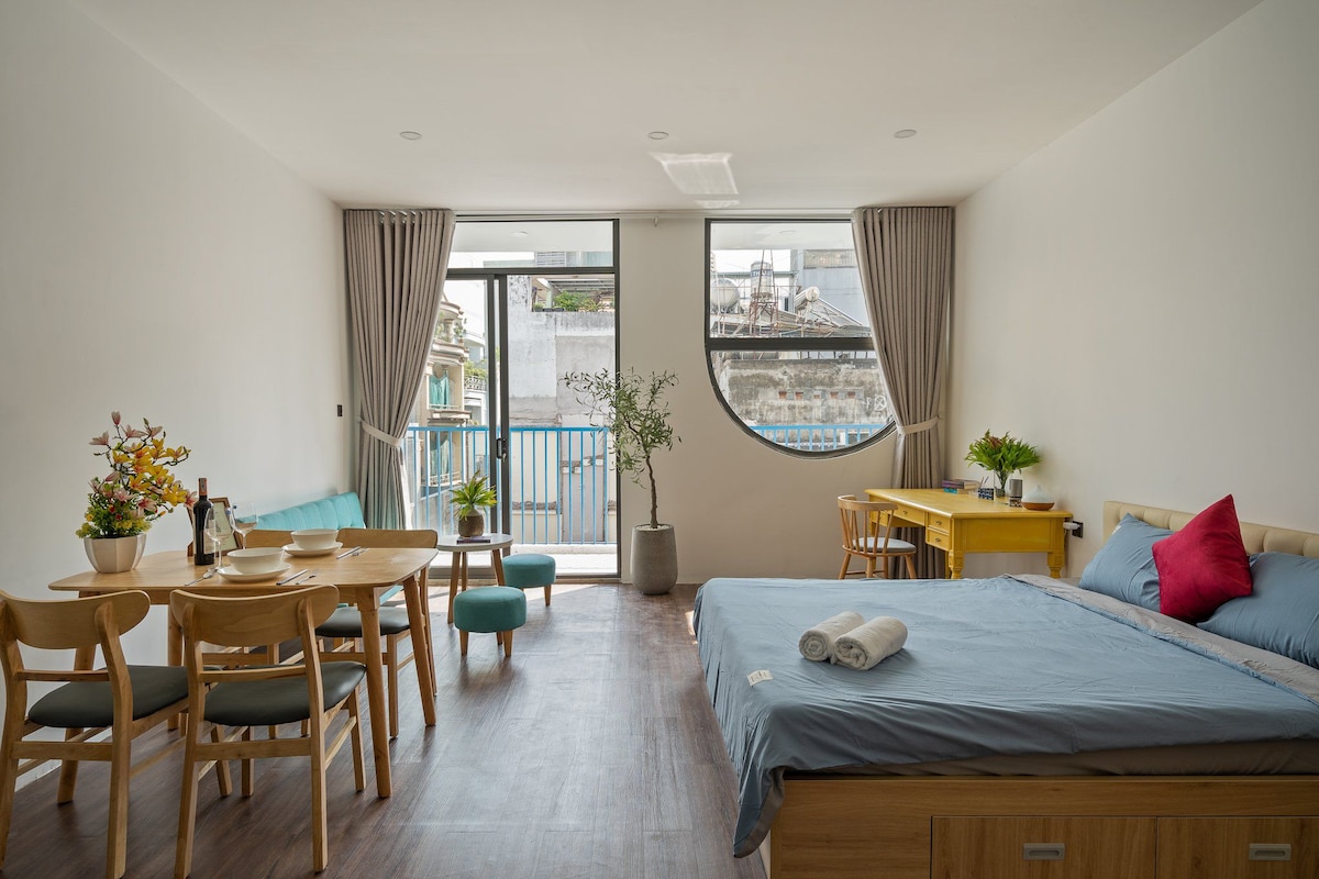 A spacious room is displayed with a comfortable sleeping area featuring a blue bedspread and accent pillows. A dining table with chairs occupies one corner, while natural light enters through large windows. Decorative plants enhance the inviting environment.