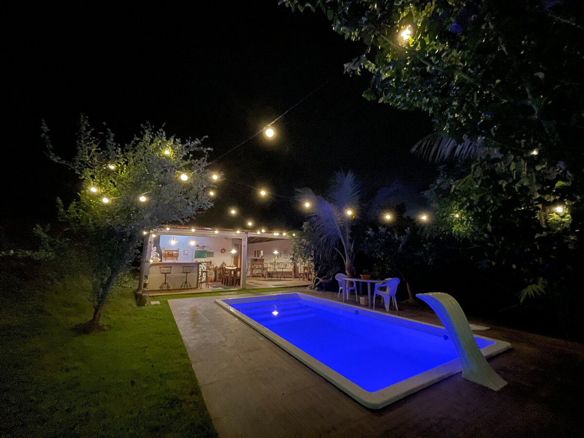 The image shows a softly illuminated pool area at night, with warm lights strung above. A bright blue pool contrasts with the surrounding greenery. Nearby, a cozy seating area features white chairs and a table, while an open space invites gatherings beneath the structure.
