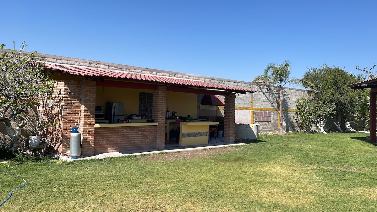 An outdoor kitchen area is featured, constructed with brick and adorned with a red roof. The space includes a countertop with appliances, surrounded by a lush green lawn and planted trees. A clear blue sky is visible above, enhancing the open atmosphere.