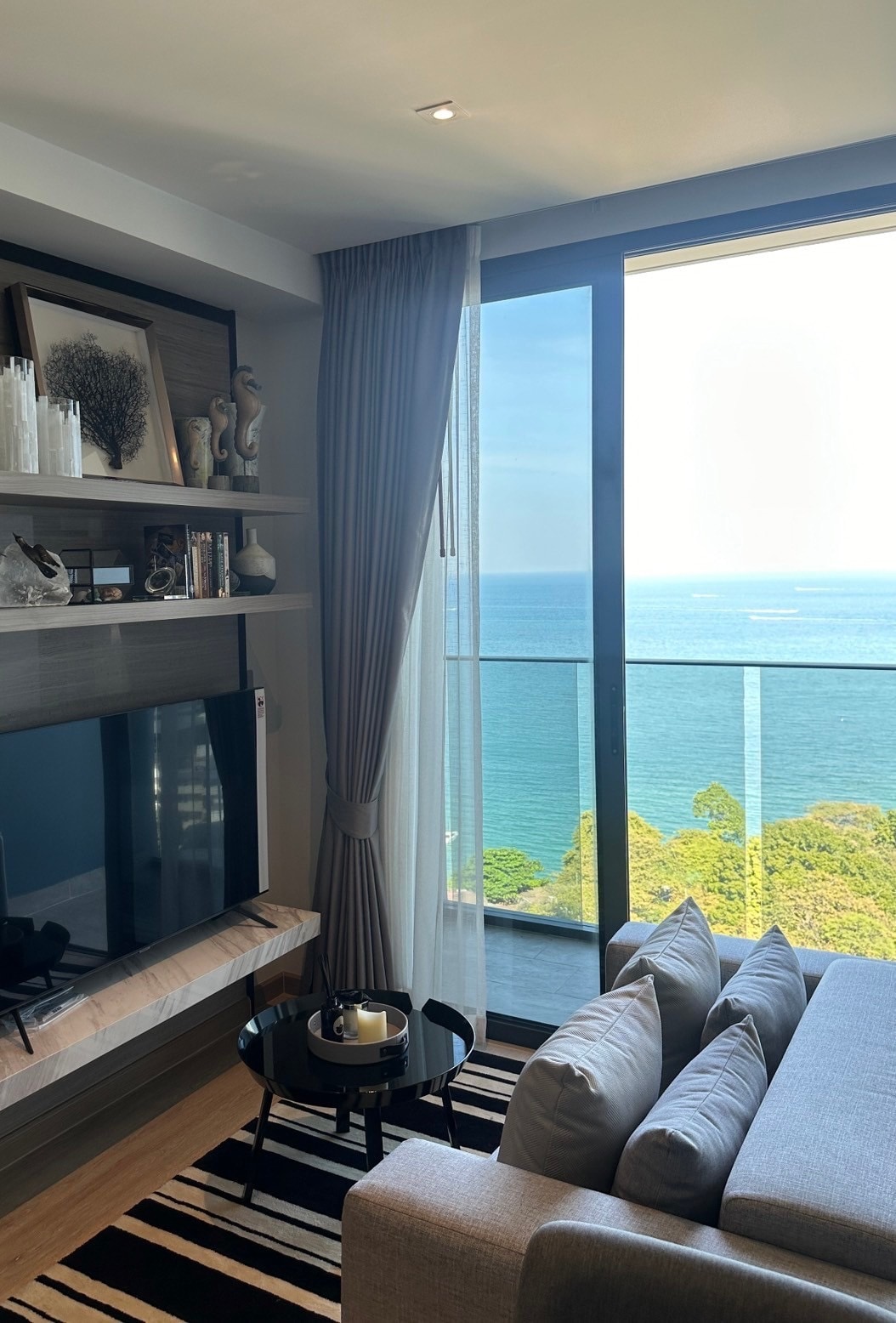 A modern living area features a large window showcasing views of the ocean. A comfortable gray sofa is positioned beside a low coffee table, while decorative shelves display art and accessories. Natural light fills the space, enhancing the open and airy atmosphere.