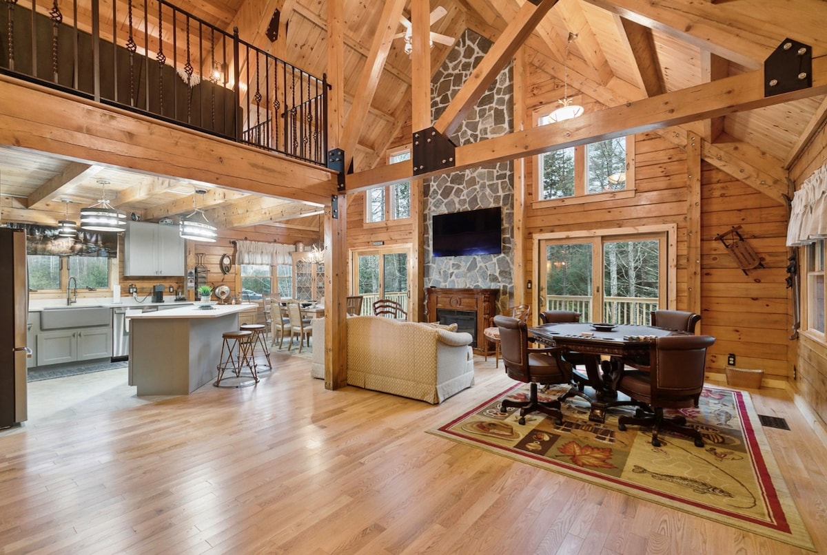 An open living space is highlighted by vaulted ceilings and wooden beams. A large stone fireplace serves as a focal point, with windows allowing natural light. A dining area with wooden chairs and a spacious kitchen are visible, alongside a comfortable seating area with a patterned rug.