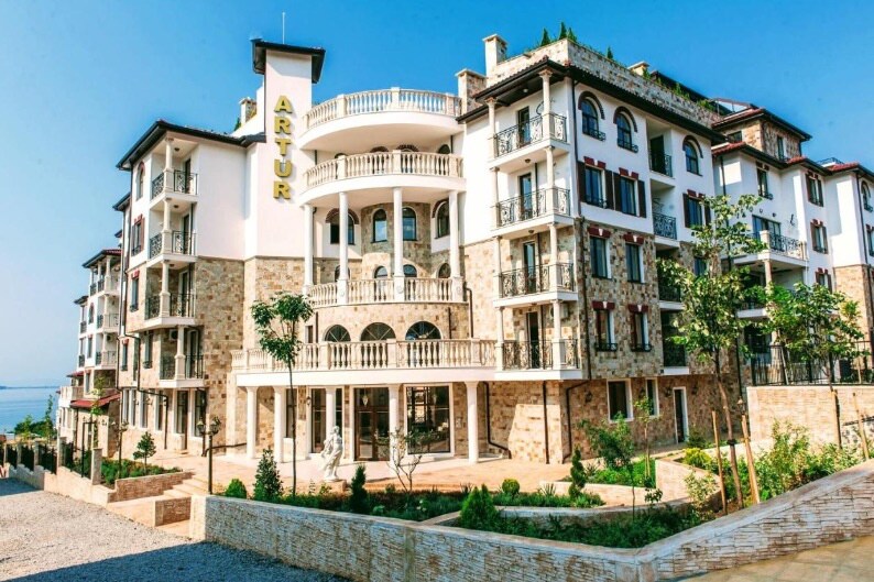 The five-story complex is characterized by a blend of stone and modern architecture. It features multiple balconies, large windows, and a welcoming entrance. Lush greenery surrounds the building, enhancing the tranquil environment near the beach.