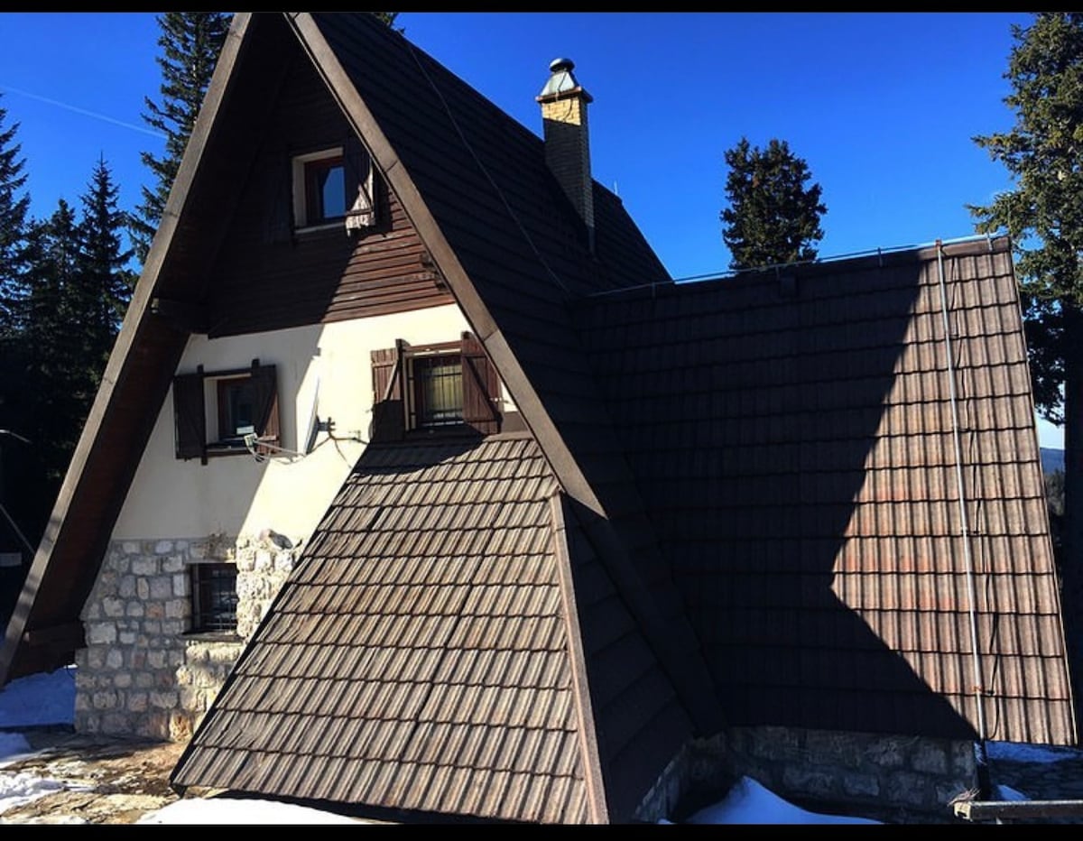 The exterior of a chalet-style house features a distinctive a-frame design with a combination of slate and wooden shingles. Sunlight casts shadows across the stone foundation, while trees stand in the background against a clear blue sky.