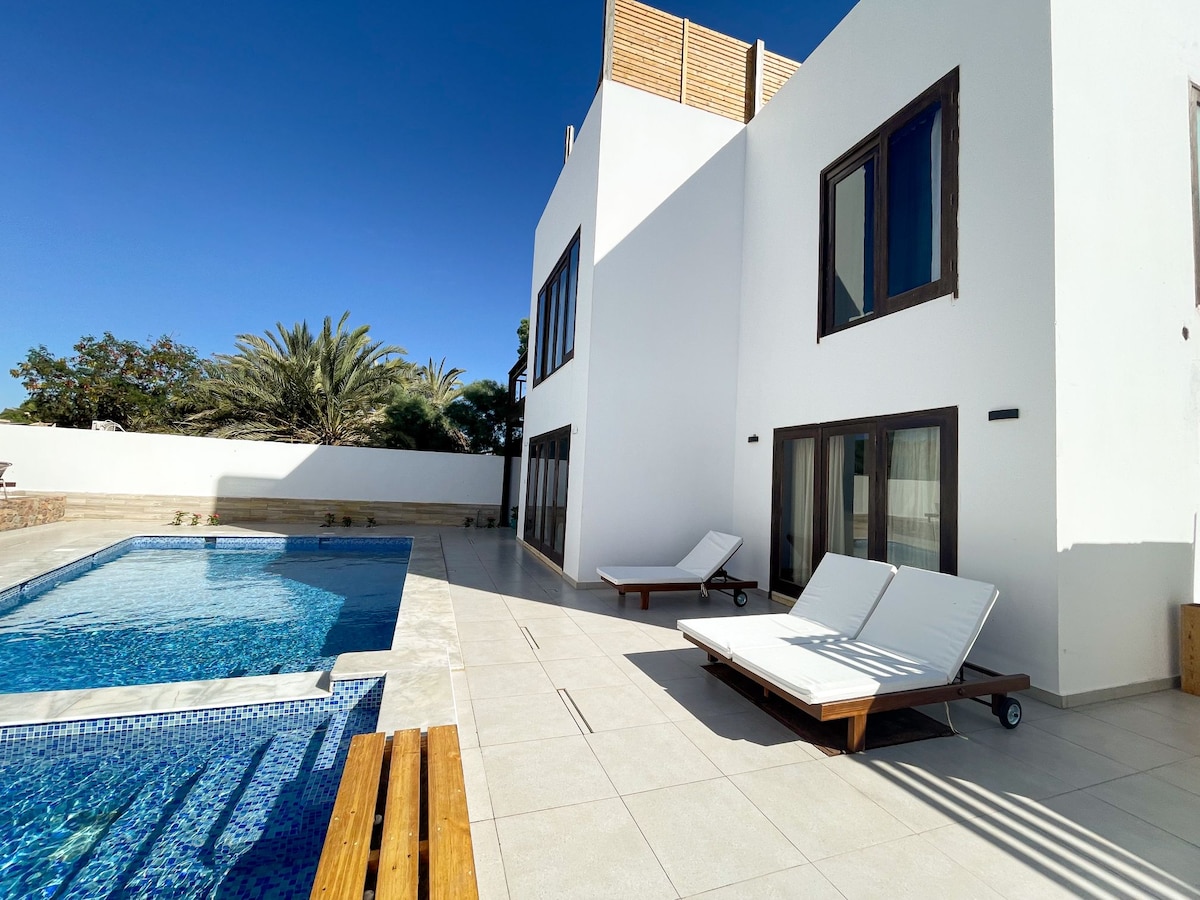 An outdoor area features a large swimming pool surrounded by a spacious terrace. Two loungers are positioned near the pool, with lush greenery visible in the background. Clear blue skies reflect the bright and open atmosphere of the villa.