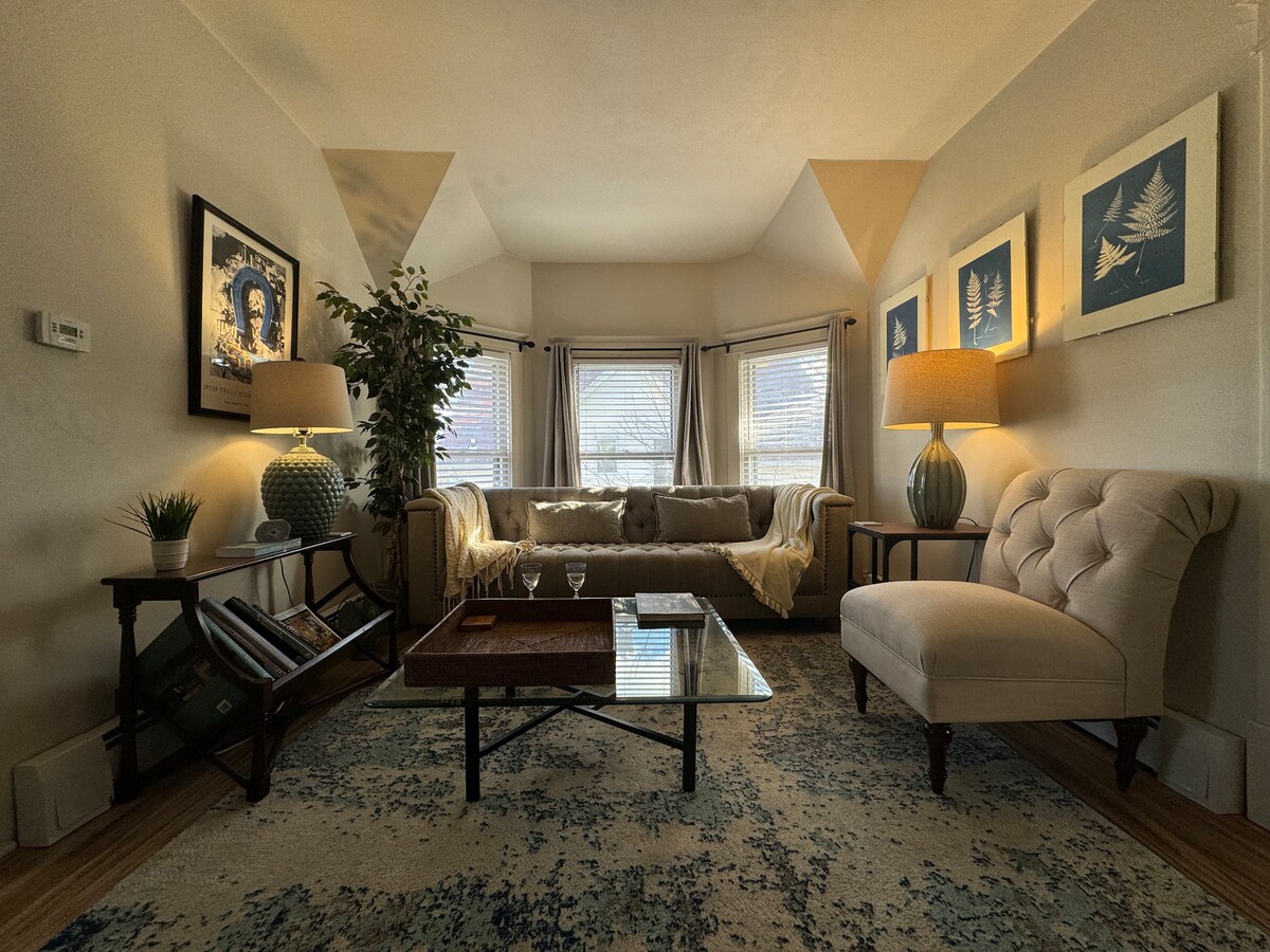 The living room features a comfortable couch accompanied by a stylish armchair and a glass coffee table. Natural light enters through the windows, illuminating framed art on the walls. A soft area rug adds warmth, while strategically placed lamps provide additional lighting.