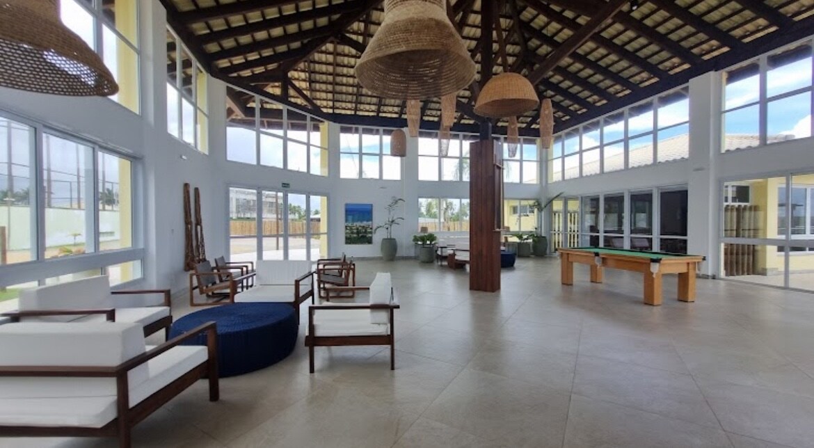 A spacious common area features large windows that allow natural light to flood the room. Comfortable seating arrangements include white chairs and a circular blue ottoman. A wooden pool table is positioned beside plants that add a touch of greenery to the space.
