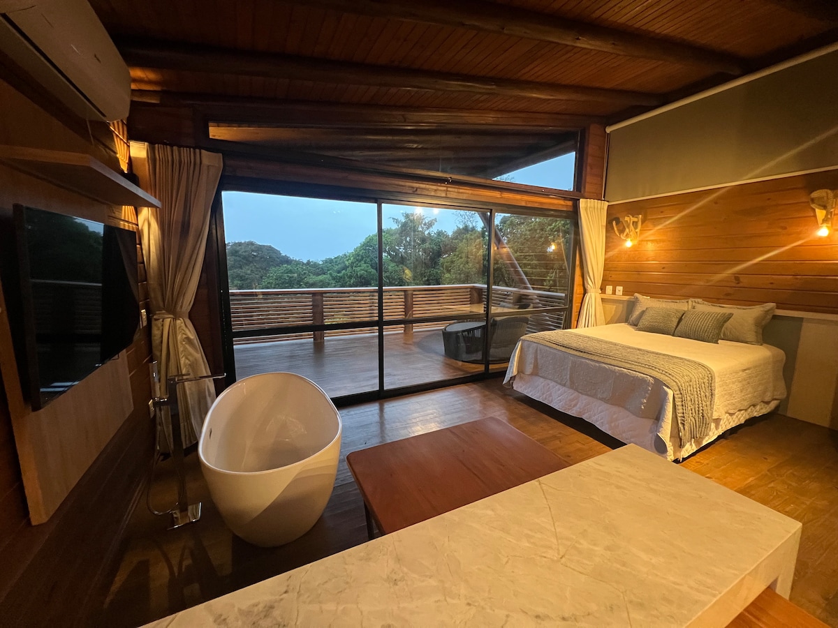 A spacious bedroom features large glass doors that open to a scenic view. A white, freestanding bathtub is positioned near the floor-to-ceiling windows, while a cozy bed adorned with soft linens is situated opposite a wooden table. Warm wood paneling complements the natural surroundings.
