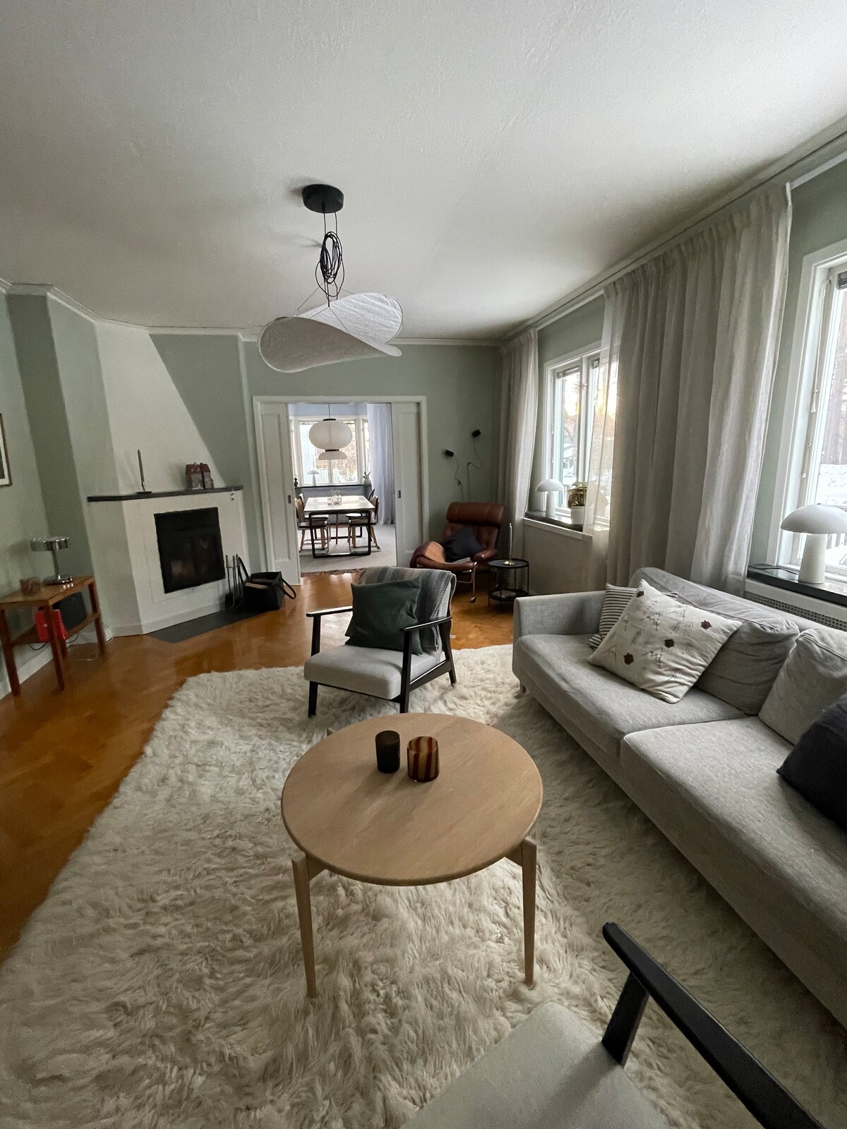A spacious living room features a soft area rug and a comfortable sofa adorned with cushions. A coffee table with decorative items sits in front, while large windows provide natural light. A fireplace adds warmth, and adjacent seating creates a cozy gathering space.