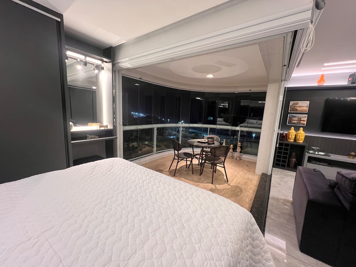 A modern room is presented, featuring a spacious bed with a quilted white coverlet. A cozy seating area with a round table and four chairs is visible beside floor-to-ceiling windows, showcasing views of the city lights. Soft ambient lighting enhances the overall atmosphere.
