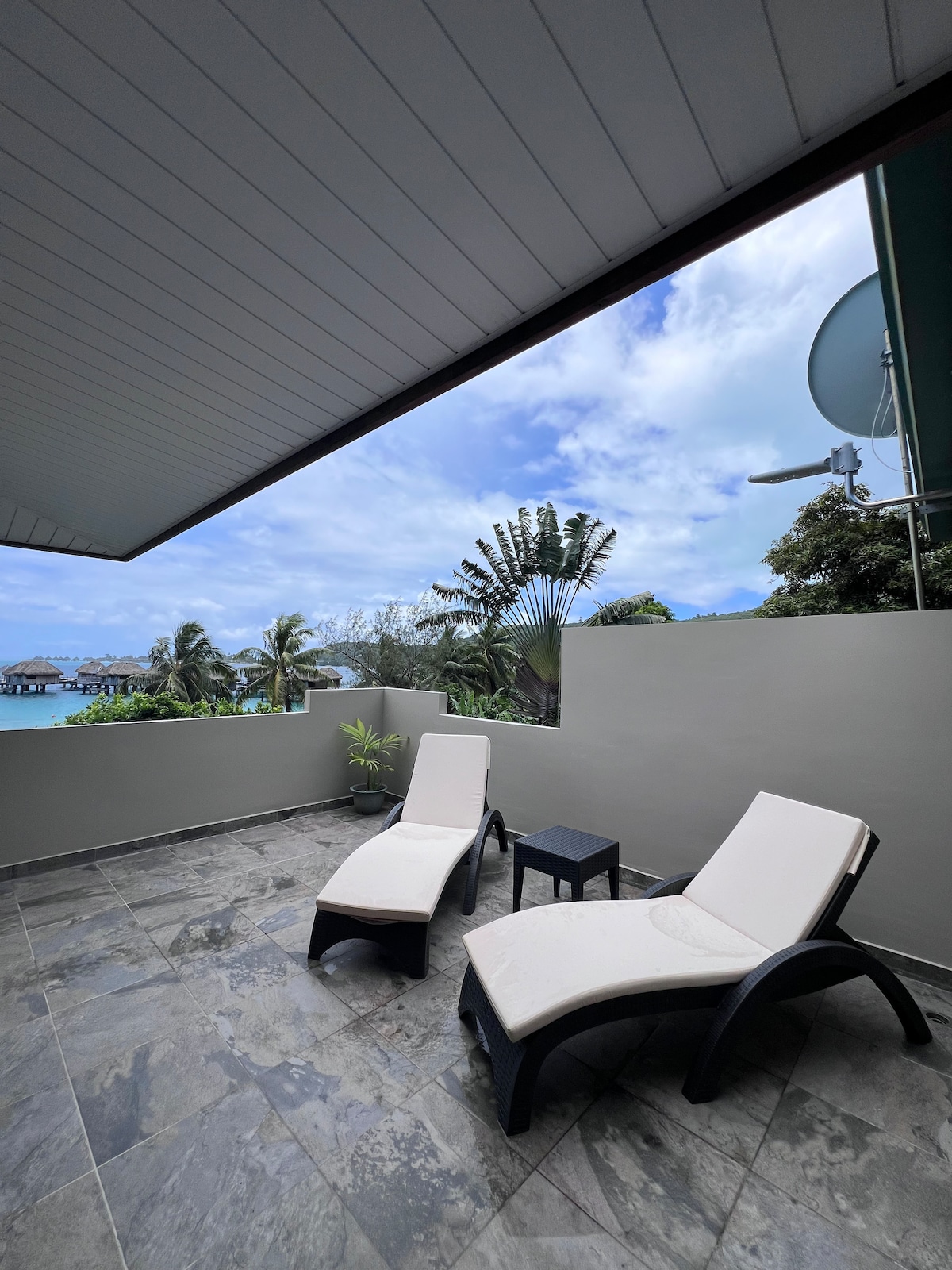 An outdoor terrace area is presented with two lounge chairs facing a view of the surrounding greenery and the lagoon. A small table sits between the chairs. The stone floor adds a cool tone, while the sky is partially cloudy.